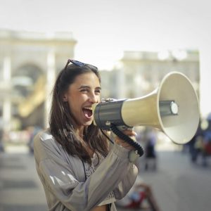 Cheerful young woman in a casual outfit shouting into a megaphone on a sunny day.